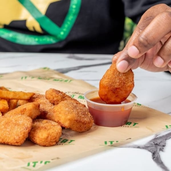 A hand dips a vegan nugget into a small cup of sauce, with fries and more nuggets nearby.
