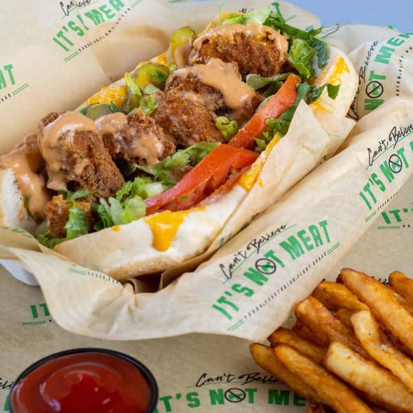 Vegan po'boy sandwich with fried plant-based protein, lettuce, tomatoes, pickles, and sauce, served with fries and ketchup.