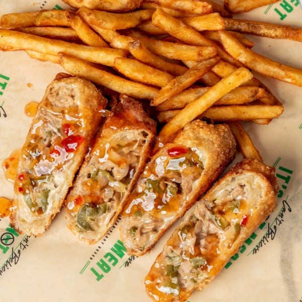Four vegan egg rolls with sweet chili sauce and a side of french fries on branded parchment paper.
